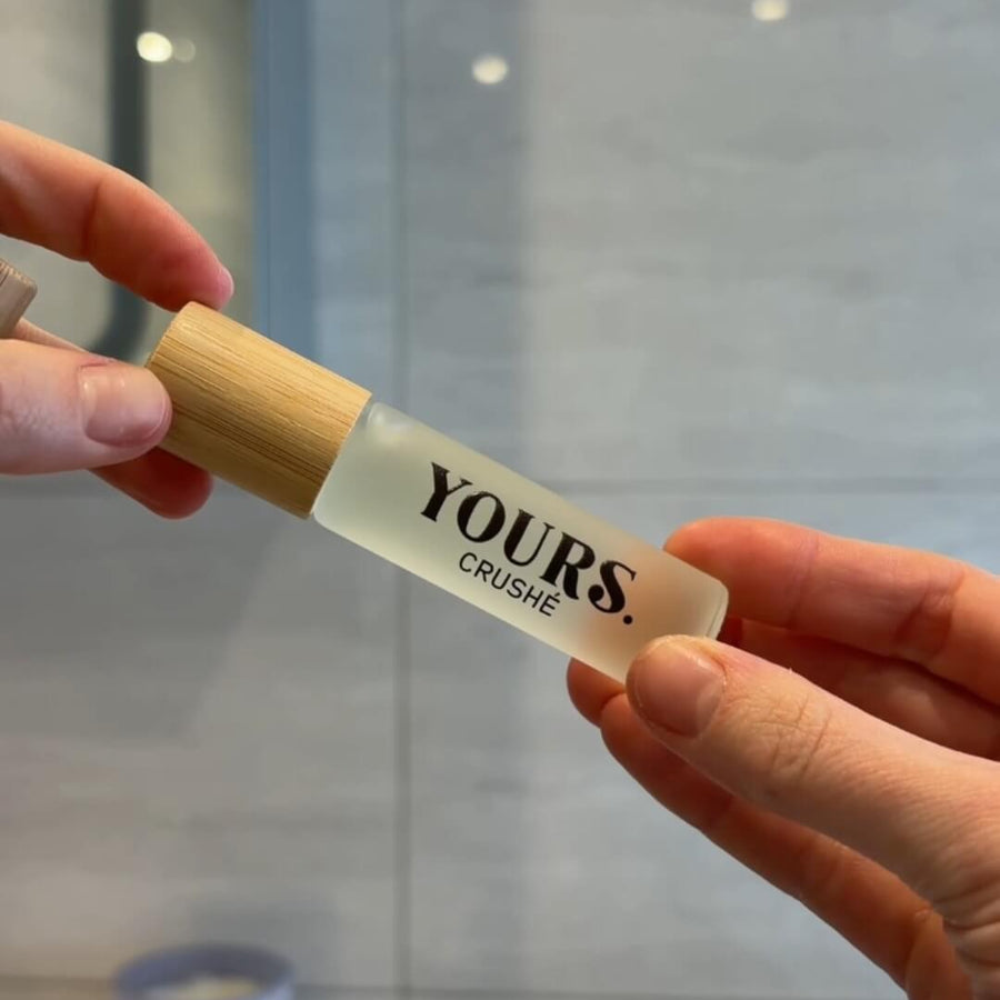 A person's hands holding a small cylindrical bottle with a light wood cap and frosted white body. The bottle is labeled 'YOURS.